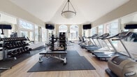 This fitness center combines elegant architectural design with a full range of exercise equipment, creating a bright and functional workout environment. The space is defined by its soaring vaulted ceiling, which peaks at a considerable height and is anchored by a decorative chain-link pendant light fixture. Large arched and rectangular windows line the walls on multiple sides, flooding the interior with natural light and offering outdoor views of the surrounding property, including a glimpse of a pool area.

The flooring features light hardwood throughout, with rubber protective mats placed beneath the weight and strength training equipment. The cardio zone is well-equipped with multiple treadmills, ellipticals, and a stationary bike, all positioned to take advantage of the expansive window views. Wall-mounted flat-screen televisions are positioned at either end of the space for entertainment during workouts.

The strength training area includes a multi-tiered barbell rack stocked with a range of weighted bars, a full dumbbell rack, an adjustable weight bench, and a cable machine. Decorative mirrored panels with geometric lattice designs and white pillar accents add a refined, upscale aesthetic to one wall, complemented by a large wall clock. The overall ambiance is airy, spacious, and polished.