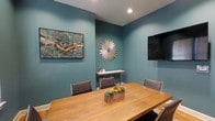 This thoughtfully designed single-floor meeting space offers a refined and professional environment suited for focused collaboration and business gatherings. The room is defined by its striking teal-painted walls, which lend a calm yet contemporary atmosphere that sets it apart from a standard conference room. A warm-toned wooden conference table serves as the centerpiece, surrounded by sleek, cushioned chairs in a neutral gray, providing both comfort and a polished aesthetic. A wall-mounted flat-screen television is positioned prominently for presentations and visual displays, ensuring functionality is at the forefront. Decorative touches elevate the space beyond the purely utilitarian — a sunburst mirror anchors the back wall with visual interest, while a framed mixed-media artwork adds texture and color to the adjacent wall. A slim, modern console table beneath the mirror offers a subtle surface for accessories or refreshments. Recessed lighting provides clean, even illumination throughout. A small wooden planter with succulents on the table introduces a natural element, softening the overall feel. Together, these details create a space that balances professional purpose with curated style.