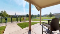 This covered outdoor pavilion is a well-appointed communal gathering space designed for comfort and functionality. Supported by sturdy structural columns beneath a gabled roof, the pavilion offers generous shelter while remaining open on all sides to embrace the surrounding landscape and natural light.

A standout feature is the built-in outdoor kitchen, constructed with attractive stacked-stone detailing and equipped with a stainless steel grill, making it ideal for outdoor cooking and entertaining. Adjacent to the kitchen, bar-height wicker seating and a round table provide a casual dining area with open views of the neighborhood greenery.

The pavilion also includes a relaxed lounge area furnished with classic Adirondack chairs and a side table, positioned to enjoy views of the lush, manicured lawn and the mature trees lining a white privacy fence. A fire pit adds an additional element for year-round outdoor enjoyment.

Looking outward from the pavilion, the views extend across well-kept green spaces, walking paths, a fenced dog park, and a trampoline area, with a landmark steeple visible on the horizon. The surrounding landscape of tall pines and open sky enhances the sense of openness and tranquility. This pavilion serves as a central outdoor amenity that combines practical features with a welcoming, park-like atmosphere.