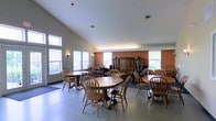 This single-floor communal space features a vaulted ceiling that creates an open, airy atmosphere throughout the room. Multiple windows along the walls allow generous natural light to fill the interior, complementing the wall sconces that provide additional ambient lighting. The space is divided into distinct functional zones: a dining area furnished with several round and rectangular oak-toned wooden tables surrounded by matching spindle-back chairs, and a lounge area toward the back equipped with upholstered seating. A prominent dark wood armoire-style cabinet serves as a visual anchor in the rear of the room, flanked by accent pieces that add a homely character. The walls feature a warm terracotta tone in portions of the space, contrasting with lighter neutral walls elsewhere to create a welcoming, residential feel. The polished concrete-style flooring is durable and easy to maintain, while a area rug near the entrance adds a touch of comfort. Overall, the space is designed to encourage social interaction in a comfortable, well-lit, and thoughtfully arranged environment.