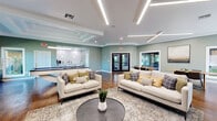 Located at 4455 SW 34th St in Gainesville, Florida, this thoughtfully designed single-floor community space combines modern aesthetics with practical functionality. Throughout the interior, rich hardwood flooring, soft sage green walls, and crisp white trim create a cohesive and inviting atmosphere.

The expansive main lounge serves as the centerpiece of the space, featuring a striking stone-surround fireplace with a natural wood mantel, a wall-mounted flat-screen TV, and stylish linear LED lighting integrated into the ceiling. Multiple seating arrangements with contemporary sofas encourage both relaxed gatherings and social interaction. A coffered ceiling detail adds architectural depth to one section of the room, while large windows and glass doors flood the interior with natural light and frame views of the surrounding lush greenery.

A dedicated game room area accommodates a full-size pool table, complementing the lounge and offering additional recreational options. The conference and co-working area features substantial natural wood tables with leather chairs, ideal for meetings or focused work sessions. Abundant windows maintain a bright, connected feel throughout.

The kitchenette is well-appointed with white cabinetry, quartz countertops, a classic subway tile backsplash, and a stainless steel sink, providing convenient refreshment access. Together, these areas deliver a versatile, welcoming environment suited for a wide range of community activities.