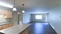 This single-floor space is designed with an open-concept layout that seamlessly connects the kitchen and living areas, creating a sense of openness and flow throughout. The kitchen features a granite countertop bar with warm wood cabinetry and is accented by two elegant pendant lights that hang from the ceiling, providing both functional and ambient lighting. The space transitions naturally into a generously sized living area finished with rich, dark hardwood flooring that runs throughout. The living area is bathed in natural light from a large window, contributing to a bright and welcoming atmosphere. A wall-mounted HVAC unit provides climate control, while a baseboard heater along the wall offers an additional source of warmth. The walls are finished in a soft, neutral gray tone that complements the overall aesthetic and creates a calm, versatile backdrop. A doorway visible from the main area leads to an additional room, suggesting further functional space beyond the open living zone. The smooth, flat ceilings and clean lines throughout give the interior a modern, understated character well-suited for a variety of uses.