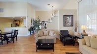This single-floor community space is designed to offer a comfortable and engaging environment for its occupants. The expansive great room serves as the heart of the space, boasting soaring vaulted ceilings that amplify the sense of openness. Large floor-to-ceiling windows fitted with vertical blinds flood the interior with abundant natural light while framing views of the surrounding exterior. Warm-toned hardwood flooring flows consistently throughout, lending a cohesive and inviting feel.

The living area is anchored by a classic fireplace topped with a wall-mounted flat-screen television, complemented by a plush sofa, loveseat, and leather accent chair arranged around a dark wood coffee table. A ceiling fan overhead adds both function and charm. Adding a touch of elegance, an upright piano is positioned near the fireplace, making the space well-suited for social gatherings and entertainment.

A full-size pool table occupies a dedicated recreation zone within the open-plan layout, while multiple round dining tables with chairs provide casual seating options bathed in natural light. A notice board and decorative indoor plants contribute to the communal atmosphere.

The kitchen features warm maple cabinetry, generous counter space, a stainless steel range with an over-the-range microwave, and a built-in sink — fully equipped to support group dining and events. A chandelier accent and framed wall art throughout add refined finishing touches to this versatile, well-appointed space.