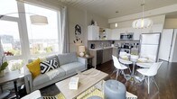 This thoughtfully designed single-floor unit seamlessly integrates the living, dining, and kitchen areas into one cohesive open-concept layout. The living area is anchored by a comfortable grey sofa accented with bold black-and-white geometric and yellow throw pillows, set atop a vibrant yellow-patterned area rug over rich dark hardwood flooring. A large floor-to-ceiling window floods the space with abundant natural light while offering impressive panoramic city views, creating a bright and airy atmosphere throughout.

The kitchen is modern and functional, featuring light wood cabinetry paired with white upper cabinets and open shelving, stainless steel appliances including a range, microwave, and dishwasher, and a clean neutral countertop workspace. A distinctive artistic wall mural adds a touch of personality and urban flair to the shared living-kitchen space.

Adjacent to the kitchen, the dining area accommodates a sleek round glass-top table surrounded by contemporary white molded chairs, ideal for casual dining. The white tongue-and-groove ceiling adds architectural character and a clean, finished look to the interior. Pendant lighting and strategically placed lamps provide warm ambient illumination, completing the polished, modern aesthetic of this well-appointed space.