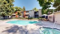 Located at 6413 Tupelo Dr in Citrus Heights, California, this single-story outdoor amenity area serves as a central recreational hub within The Woods community. The space is anchored by a large, rectangular swimming pool with clear, blue-tiled water, surrounded by an expansive concrete pool deck ideal for sunbathing and lounging. Blue lounge chairs are positioned along the deck, providing comfortable spaces to relax in the California sun.

Adjacent to the main pool is a separate octagonal spa, offering a more intimate aquatic retreat. Bright red French doors lead into an interior clubhouse or common area, connecting the indoor and outdoor spaces seamlessly. An outdoor seating area with a table, chairs, and a blue umbrella provides a shaded spot for dining or socializing poolside.

A utility building along the perimeter houses multiple private changing rooms or restroom facilities, each with individual doors, ensuring convenience for guests. The entire area is enclosed by a wrought iron fence, providing security and privacy. Mature evergreen and deciduous trees surround the space, creating a naturally shaded and serene atmosphere. The lush greenery and landscaped planters further enhance the park-like setting, making this outdoor amenity area a welcoming retreat for residents.