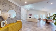 This leasing office in Walls, Mississippi, is a thoughtfully designed single-story space that balances professional functionality with upscale interior aesthetics. The layout flows seamlessly across several distinct zones, all unified by wide-plank wood-look flooring, crisp white walls, and elegant crown molding.

The reception area makes an immediate impression with its dramatic vaulted ceiling, a bold 3D geometric accent wall in gray, and a gold textured credenza topped with a large round mirror. A cascading circular chandelier anchors the space overhead, while recessed lighting supplements the natural light streaming through multiple windows with plantation shutters.

The lounge area features a plush white sectional sofa atop a marble-patterned area rug, a wall-mounted flat-screen TV above a sleek electric fireplace console, and a statement gold bubble chair — adding a playful, modern focal point. French doors throughout invite natural light and outdoor views, enhancing the open, airy atmosphere.

The workstation area transitions smoothly from the lounge and includes multiple staff desks illuminated by circular pendant lights, accompanied by upholstered guest chairs in warm blush and taupe tones. A separate consultation room with a round table, pink velvet chairs, and abundant window light provides a welcoming space for private meetings. The overall ambiance is polished, contemporary, and inviting.