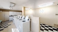 This single-floor laundry facility is designed to accommodate multiple users simultaneously, offering a practical and functional layout. The room features a notable black-and-white checkered tile floor that lends a classic, retro aesthetic to the space. Vertical wood panel wainscoting in a cream tone lines the walls, contributing to a clean, uniform appearance throughout.

The facility is equipped with a row of top-loading washing machines along one wall, complemented by a bank of stacked dryers positioned nearby, providing ample capacity for residents. Extended, flat folding counters run along the center and perimeter of the room, offering generous surface space for sorting and folding laundry. A small seating area with light teal chairs is available for those waiting during wash cycles.

Natural light filters in through a series of horizontal windows fitted with security bars, positioned along the upper portion of the walls. Overhead fluorescent lighting ensures the space remains well-lit at all times. A storage cabinet is also present, adding to the room's overall utility. Posted signage and bulletin materials are visible, keeping users informed of facility rules and instructions.