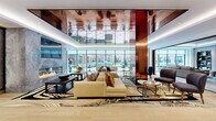 This upscale amenity space is a masterclass in contemporary interior design, thoughtfully composed across a single floor with distinct yet seamlessly connected lounge areas. The centerpiece of the space is a dramatic, polished copper-toned reflective ceiling that crowns the main lounge, adding warmth and visual depth. Floor-to-ceiling glass walls and large picture windows flood the interior with natural light while offering expansive views of a landscaped outdoor courtyard, creating a strong indoor-outdoor connection. The main lounge is anchored by a sleek, linear gas fireplace set against a richly textured stone wall, complemented by generous sectional sofas and bold, patterned area rugs that lend an artistic flair. Gold-accented side tables and carefully chosen accent pillows in earthy tones add refined detail throughout. A secondary lounge area features warm wood-paneled ceilings with recessed ambient lighting, additional plush sectional seating, and continued views of the courtyard beyond. A standout architectural feature is the glass-enclosed wine display room, framed by matte black steel doors, lit with decorative globe pendants, and showcasing an impressive built-in wine rack. Light hardwood flooring unifies the space, contributing to its clean, modern aesthetic. The overall ambiance is polished, inviting, and distinctly upscale.