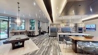 This expansive single-floor amenity space is designed with a contemporary aesthetic that seamlessly integrates multiple functional zones into one cohesive environment. Throughout the space, large-format porcelain tile flooring in variegated grey and wood tones creates a refined, continuous visual foundation.

The fully equipped kitchen serves as a centerpiece, featuring professional-grade stainless steel appliances, a large marble-topped island with seating for several, sage-toned cabinetry with subway tile backsplash, and a striking pair of geometric gold pendant lights overhead. A large stainless range hood anchors the cooking area.

Adjacent to the kitchen, the entertainment zone accommodates a full-size billiards table beneath sculptural pendant lighting, alongside a wall-mounted display and decorative mirrored panels. A built-in bar counter with high seating faces floor-to-ceiling windows that frame views of a lush outdoor pool area.

The lounge areas offer comfortable sectional seating beneath tray ceilings with warm cove lighting, while a quiet sitting nook displays curated artwork and accent chairs. Throughout, dark wood millwork, recessed lighting, and large windows contribute to a polished, welcoming ambiance.