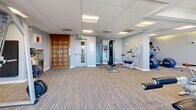 Located at 100 Steele St in Denver's Cherry Creek neighborhood, this fitness center offers a comprehensive workout environment across a single, open floor plan. The space is finished with durable striped carpeting throughout and features a suspended drop ceiling with recessed and panel lighting that keeps the facility bright and well-lit.

The facility is thoughtfully divided into functional workout zones. A dedicated cardio area houses treadmills, ellipticals, stationary bikes, and rowing machines, positioned near floor-to-ceiling sliding glass doors that open to an outdoor balcony. This connection to the exterior allows for abundant natural light and sweeping views of the Rocky Mountains, creating an energizing atmosphere during workouts.

The strength training section is well-appointed with a full rack of dumbbells, medicine balls, resistance bands, and a multi-use cable and weight machine. A barbell rack with plate weights supports heavier lifting, while wall-mounted mirrors enhance the sense of space and allow for form checks. Additional cardio and low-impact equipment, including stability balls and recumbent options, are housed in a quieter, secondary zone with softer ambient lighting. A warm wood shelving unit near the entrance provides storage, and the facility is accessible via a secured entry door with clear signage throughout.