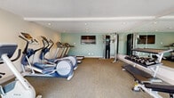 This single-level fitness center in Alameda, California, offers a well-organized and efficiently designed workout space. The room features neutral-toned walls accented by a soft aqua-blue feature wall, complemented by carpet flooring throughout and recessed lighting in the white ceiling, creating a clean and functional atmosphere.

The cardio zone is equipped with multiple treadmills and elliptical trainers, arranged to face a wall-mounted flat-screen television, making workouts engaging and convenient. A full cable multi-use strength training station is positioned nearby, accompanied by a dumbbell rack stocked with a range of free weights and a dedicated weight bench, providing a comprehensive strength training area.

Along one side of the room, a large mirrored wall enhances the sense of openness and allows users to monitor their form during exercise. On the opposite end, large windows and a glass door flood the space with natural light and offer views of a landscaped outdoor area. A wall-mounted accessory storage rack holds medicine balls, resistance equipment, and additional fitness accessories. A split-unit air conditioning system ensures year-round climate comfort. The overall layout is practical and uncluttered, making effective use of the available footprint.