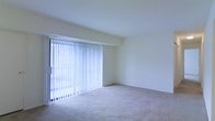 This single-floor space presents a clean, minimalist living area characterized by its bright and airy atmosphere. The room is finished with neutral-toned wall-to-wall carpeting that adds warmth and comfort underfoot, complementing the crisp white walls throughout. A prominent feature of the space is the large sliding glass door fitted with full-length vertical blinds, which serves as the primary source of natural light and provides access to the outdoors. The blinds offer flexible light control while maintaining a sense of openness. The walls are smooth and freshly painted in a light, neutral tone, creating a versatile backdrop suitable for a variety of interior styles. The layout is straightforward and open, allowing for flexible furniture arrangement. Subtle architectural detailing includes a gentle transition between the wall and ceiling, adding modest visual interest to the otherwise understated design. The overall ambiance is clean, quiet, and functional — well-suited for those seeking a simple, move-in-ready living environment with an emphasis on natural light and uncluttered space.