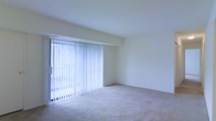 This single-floor space presents a clean, minimalist living area characterized by its bright and airy atmosphere. The room is finished with neutral-toned wall-to-wall carpeting that adds warmth and comfort underfoot, complementing the crisp white walls throughout. A prominent feature of the space is the large sliding glass door fitted with full-length vertical blinds, which serves as the primary source of natural light and provides access to the outdoors. The blinds offer flexible light control while maintaining a sense of openness. The walls are smooth and freshly painted in a light, neutral tone, creating a versatile backdrop suitable for a variety of interior styles. The layout is straightforward and open, allowing for flexible furniture arrangement. Subtle architectural detailing includes a gentle transition between the wall and ceiling, adding modest visual interest to the otherwise understated design. The overall ambiance is clean, quiet, and functional — well-suited for those seeking a simple, move-in-ready living environment with an emphasis on natural light and uncluttered space.