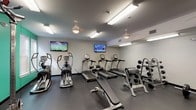 Located at 10300 University Parkway in Pensacola, Florida, this well-equipped fitness center is designed to support a full range of workout routines in a single-floor layout. The space is divided into distinct functional zones, each thoughtfully outfitted with professional-grade equipment.

The cardio area features multiple treadmills, elliptical machines, and a stationary bike, all positioned beneath ceiling fans and bright fluorescent lighting for a comfortable workout environment. Wall-mounted flat-screen televisions are strategically placed throughout, keeping users entertained during cardio sessions.

The free weights section includes a comprehensive dumbbell rack with a wide range of weights, a barbell with plates, and a flat bench, complemented by large wall mirrors that enhance the sense of space and allow for proper form checks. A dedicated strength training zone houses a multi-functional cable machine, a squat rack with an Olympic barbell, and additional benching options — catering to both compound and isolated strength movements.

The facility features durable rubber flooring throughout, providing safety and sound absorption across all workout zones. Accent walls in bold blue and green add a vibrant, energizing atmosphere to the space. Ceiling fans and overhead lighting ensure consistent airflow and visibility. The fitness center is accessible via a dedicated entry door, making it a convenient and complete amenity.