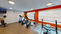 Located at 25 Landis Ln in Lillington, North Carolina, this single-floor fitness center is designed to support a well-rounded workout routine in a vibrant, purpose-built environment. The space is immediately defined by its bold orange geometric accent wall, which adds an energizing visual contrast against the neutral gray walls and textured ceiling. Durable, speckled rubber flooring covers the entire area, providing a practical and resilient surface suited for a range of exercises. Natural light streams through multiple windows fitted with white blinds, complementing the flush-mount ceiling lights that ensure consistent, bright illumination throughout. The cardio zone is well-equipped with a treadmill, elliptical, and stationary bike, all arranged to take advantage of the natural light and a wall-mounted flat-screen television. The free weights area features a multi-tier dumbbell rack stocked with a variety of weights, along with barbells and weight plates, a weight bench, and a squat rack. A medicine ball storage rack and a standing floor fan round out the functional amenities. Full-length mirrors along one wall expand the visual space and support proper form during training. A posted gym etiquette sign reflects the space's commitment to a respectful fitness environment.
