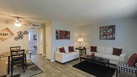 This single-floor space offers a comfortable and functional layout centered around a well-lit living area. The living room features wood-style plank flooring that extends throughout the main area, providing a warm and cohesive aesthetic. Neutral-toned walls in soft white and beige create a clean backdrop, complemented by a textured popcorn ceiling. The space is furnished with white upholstered sofas accented by deep red throw pillows, anchored by a dark coffee table and area rug, lending a contemporary contrast to the light surroundings. Decorative wall art adds personality and visual interest to the space. A central hallway connects the living area to the rest of the unit, with a glimpse into a carpeted bedroom visible in the background, suggesting a clear and practical flow between rooms. Overhead HVAC vents are integrated into the ceiling, indicating a centralized climate control system. A floor lamp provides warm ambient lighting, enhancing the inviting atmosphere. Overall, the space presents a straightforward, move-in-ready layout suited for everyday living.