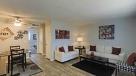 This single-floor space offers a comfortable and functional layout centered around a well-lit living area. The living room features wood-style plank flooring that extends throughout the main area, providing a warm and cohesive aesthetic. Neutral-toned walls in soft white and beige create a clean backdrop, complemented by a textured popcorn ceiling. The space is furnished with white upholstered sofas accented by deep red throw pillows, anchored by a dark coffee table and area rug, lending a contemporary contrast to the light surroundings. Decorative wall art adds personality and visual interest to the space. A central hallway connects the living area to the rest of the unit, with a glimpse into a carpeted bedroom visible in the background, suggesting a clear and practical flow between rooms. Overhead HVAC vents are integrated into the ceiling, indicating a centralized climate control system. A floor lamp provides warm ambient lighting, enhancing the inviting atmosphere. Overall, the space presents a straightforward, move-in-ready layout suited for everyday living.