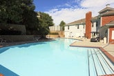 This single-level outdoor amenity space offers a well-rounded recreational environment designed for community use. The centerpiece is a generously sized swimming pool with a classic brick-edged surround and a dedicated diving board platform, providing ample space for both leisurely and active use. The pool deck is finished in smooth concrete and lined with multiple lounge chairs, creating a comfortable sunbathing area shaded in part by mature trees that border the space.

Adjacent to the pool area stands a covered octagonal gazebo with a warm wood-paneled ceiling and a ceiling fan, offering a shaded retreat with outdoor seating. The gazebo overlooks the surrounding green space and provides views toward a children's playground equipped with a slide and climbing structure, enclosed within a secure fenced perimeter.

The grounds are thoughtfully landscaped with trimmed hedges, ornamental shrubs, flowering plants, and tiered retaining walls clad in brick, adding structure and visual interest. Wrought iron fencing defines the boundaries of the space, contributing to both security and aesthetic cohesion. A brick accessory building is positioned poolside, likely serving as a utility or restroom facility. The overall environment is bright, open, and well-maintained under natural daylight.