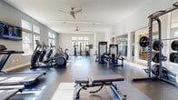 Located at 100 Forge Circle in Lawrence Township, NJ, this well-appointed fitness center offers a generously sized, single-floor layout designed to accommodate a comprehensive range of workout activities. The facility features large windows along multiple walls, flooding the space with natural light and providing views of the surrounding outdoor environment. Ceiling fans are mounted overhead, contributing to air circulation and comfort throughout the room.

The flooring consists of large-format dark tiles with cushioned mat sections strategically placed beneath equipment zones. The cardio area is equipped with multiple treadmills, elliptical trainers, stair climbers, and stationary bikes, all positioned to take advantage of the natural light and wall-mounted televisions. The strength training section includes a variety of cable and resistance machines, adjustable benches, and a dedicated free weight area with a full dumbbell rack.

A motivational athletic mural with bold red and black graphic design accents one wall, adding visual energy to the space. Glass partitions along one side provide a sense of openness and connectivity. The overall ambiance is clean, modern, and bright, making this fitness center a fully equipped destination for both cardio and strength training needs.