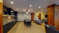 This well-appointed single-floor space showcases a thoughtfully designed open-plan layout that seamlessly integrates a fully equipped kitchen, a formal dining area, and a spacious living zone. Light hardwood flooring runs continuously throughout, providing a warm and cohesive foundation for the interior.

The kitchen is a standout feature, outfitted with sleek navy cabinetry, a gold vertical-tile mosaic backsplash, stainless steel appliances including a built-in oven and cooktop, and open shelving with under-cabinet lighting. Warm walnut wall panels frame the kitchen, adding visual depth and contrast.

The dining area centers around a long dark table with seating for several guests, illuminated by a decorative crystal chandelier. Adjacent, the living area is generously proportioned, furnished with a large sectional sofa, accent chairs, a mounted flat-screen television, and a low-profile media console.

Tall walnut-clad structural columns define zones throughout the space while reinforcing the warm, contemporary aesthetic. A dramatic floor-to-ceiling glazed window wall floods the interior with natural light and offers views of a landscaped outdoor terrace. Sheer linen curtains soften the light while maintaining openness. Curated artwork is displayed throughout, completing the polished, design-forward ambiance.
