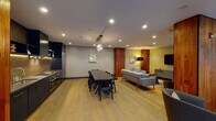 This well-appointed single-floor space showcases a thoughtfully designed open-plan layout that seamlessly integrates a fully equipped kitchen, a formal dining area, and a spacious living zone. Light hardwood flooring runs continuously throughout, providing a warm and cohesive foundation for the interior.

The kitchen is a standout feature, outfitted with sleek navy cabinetry, a gold vertical-tile mosaic backsplash, stainless steel appliances including a built-in oven and cooktop, and open shelving with under-cabinet lighting. Warm walnut wall panels frame the kitchen, adding visual depth and contrast.

The dining area centers around a long dark table with seating for several guests, illuminated by a decorative crystal chandelier. Adjacent, the living area is generously proportioned, furnished with a large sectional sofa, accent chairs, a mounted flat-screen television, and a low-profile media console.

Tall walnut-clad structural columns define zones throughout the space while reinforcing the warm, contemporary aesthetic. A dramatic floor-to-ceiling glazed window wall floods the interior with natural light and offers views of a landscaped outdoor terrace. Sheer linen curtains soften the light while maintaining openness. Curated artwork is displayed throughout, completing the polished, design-forward ambiance.