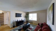 This single-floor space centers around a well-appointed living room that balances functionality with a welcoming ambiance. The room features warm hardwood flooring that extends throughout, complemented by neutral-toned walls that create a clean, versatile backdrop. A textured popcorn ceiling adds a classic touch overhead.

The living area is anchored by a wall-mounted flat-screen television positioned above a sleek black media console, which provides ample storage with multiple drawers and shelving. Decorative accents, including tall vases and ornamental grasses, are arranged atop the console, adding visual interest. A deep burgundy sofa with patterned throw pillows anchors the seating area, paired with an oval upholstered ottoman serving as a central coffee table. A geometric patterned area rug defines the seating zone.

Natural light enters through a large window dressed with white horizontal blinds, brightening the space and offering a sense of openness. A tall indoor palm plant near the window introduces a touch of greenery. A white interior door is positioned adjacent to the window, contributing to the room's clean, organized layout.