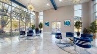 This contemporary community clubhouse is designed to impress from the moment you step inside. The grand entrance lobby commands attention with soaring double-height ceilings, an expansive floor-to-ceiling window wall that floods the space with natural light, and a striking starburst gold chandelier that serves as a stunning focal point. Large-format hexagonal floor tiles in a soft neutral tone run throughout the entire facility, creating a cohesive and polished aesthetic. Royal blue velvet accent chairs, arranged in intimate seating clusters atop layered rugs, lend a bold, modern character to the welcoming lobby. Abstract blue-toned artwork adorns the walls, reinforcing the space's vibrant color palette. Moving deeper into the clubhouse, a spacious lounge area features curved navy blue sofas arranged around a tile-surround fireplace, with views of the outdoor pool area through wide windows. A well-appointed kitchenette offers white cabinetry, stone countertops, a full-length bar with seating, and a textured tile backsplash. The game room rounds out the amenities with a billiards table, high-top bar seating, casual dining tables, and open sightlines connecting all areas of the floor. Lush tropical plants are placed throughout, adding warmth and life to this bright, well-curated communal space.