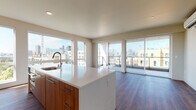 This contemporary single-floor residence showcases a thoughtfully designed open-concept layout with rich hardwood flooring throughout. The kitchen serves as the centerpiece of the living space, anchored by a generous island with a striking white quartz waterfall countertop and an undermount sink. Warm wood cabinetry lines the walls with under-cabinet lighting illuminating a marble-style backsplash, complemented by stainless steel appliances including a gas range. Large floor-to-ceiling windows and sliding glass doors flood the living area with abundant natural light and frame sweeping city skyline views, opening directly onto a private balcony. The primary bedroom features its own sliding glass door to the balcony and is accompanied by a spacious walk-in closet outfitted with adjustable wire shelving systems. The primary bathroom offers a glass-enclosed walk-in shower with large-format linear tile and a mosaic tile floor. A secondary bathroom includes a full soaking tub with matching tile surround and chrome fixtures. A dedicated laundry area houses a stacked washer and dryer unit alongside additional closet storage. The overall design is clean, modern, and cohesive, with a neutral palette that enhances the sense of openness throughout.