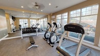 This single-floor fitness center offers a thoughtfully designed workout environment that balances functionality with a bright, welcoming atmosphere. Large, multi-pane windows line the walls, flooding the space with natural light and providing open views of the surrounding outdoor greenery, creating an energizing backdrop for exercise.

The facility is outfitted with a comprehensive selection of True-brand cardio equipment, including treadmills, elliptical trainers, and a stationary bike, catering to a variety of aerobic training preferences. A well-organized free weight station features a full rack of dumbbells alongside an adjustable weight bench, accommodating both beginner and advanced strength training routines.

The interior finishes are clean and modern, with neutral-toned walls accented by bold horizontal stripes of orange and gray in one section, adding a dynamic visual element to the space. Carpet tile flooring throughout provides comfort underfoot during workouts. A large wall-mounted mirror expands the sense of space and allows users to monitor their form. Ceiling fans with integrated lighting fixtures are distributed throughout, ensuring consistent airflow and comfort. Recessed lighting supplements the natural light, maintaining excellent visibility at all hours. A glass-paneled exterior door offers convenient direct access to the outdoor area.