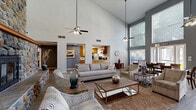 Located at 2593 Millcreek Dr in Sacramento, CA, this single-floor community space offers an impressive and open layout designed for comfort and gathering. The living room is defined by its dramatic double-height ceilings and a striking floor-to-ceiling river rock fireplace with a warm wood mantle, serving as the room's undeniable focal point. Durable wood-look tile flooring runs throughout, complementing the neutral gray and white wall tones. A ceiling fan provides additional comfort, while the expansive wall space accommodates an entertainment center and seating area, creating an inviting atmosphere for relaxation. The dining area flows naturally from the living space, featuring large windows with horizontal blinds that allow abundant natural light to pour in and offer views of the surrounding greenery. A generously sized wooden dining table with ample seating makes the area well-suited for group gatherings. Direct exterior access via a glass-paneled door further enhances the connection to the outdoors. Adjacent to the dining room, the kitchen is functional and well-appointed, featuring warm honey-toned wood cabinetry, granite-style countertops, a stainless steel refrigerator, and tile flooring. Ample upper and lower cabinet storage ensures strong utility throughout. Together, these spaces form a cohesive, light-filled environment ideal for community use.
