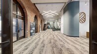 Located at 1000 Washington Ave in downtown St. Louis, this expansive single-floor amenity space masterfully blends historic architecture with contemporary design. The building's original exposed brick walls, dramatic arched openings, and soaring ceilings create a striking industrial backdrop, complemented by modern finishes and carefully curated furnishings throughout.

The entrance greets visitors through glass-paneled doors set within a stone façade, opening into a long, light-filled corridor featuring geometric LED lighting and patterned carpet tile flooring. The main gathering area is an open, airy lounge anchored by white structural columns and exposed ductwork overhead. Seating arrangements include curved green sofas, leather couches, and accent chairs, alongside a sleek dark fireplace feature wall that serves as a focal point.

A dedicated lounge area near floor-to-ceiling street-facing windows floods the space with natural light and offers views of Washington Avenue. Bar-height seating and café tables line the windows, providing casual dining and workspace options. A game room features a shuffleboard table set against the original brick wall. A staffed leasing or concierge desk with a branded wood logo medallion anchors one end of the floor. Multiple distinct seating vignettes offer varied atmospheres for socializing, working, or relaxing throughout this generously proportioned space.