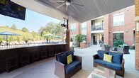 Located at 260 Leigh Farm Rd in Durham, NC, this outdoor amenity space is designed to deliver a resort-style experience within a well-appointed residential community. The centerpiece is a generously sized swimming pool with a shallow sun shelf lined with in-water lounge chairs, surrounded by an expansive pool deck furnished with chaise lounges and shaded by colorful umbrellas — ideal for relaxing under the North Carolina sun.

Adjacent to the pool, a covered outdoor lounge area features sturdy brick columns supporting a vaulted ceiling equipped with ceiling fans, mounted flat-screen televisions, and overhead heaters, ensuring year-round usability. Comfortable wicker seating with navy blue and yellow accent cushions is arranged throughout, creating inviting conversation areas. A built-in outdoor bar with bar-stool seating sits within the covered zone, directly overlooking the pool area.

Beyond the lounge, the open-air patio extends to include dedicated grilling stations with stainless steel gas grills built into brick surrounds, along with a striking brick fireplace feature for evening gatherings. Lush landscaping, manicured evergreens, and architectural lighting enhance the courtyard setting. The multi-story brick and stucco building rises around the space, with resident balconies offering elevated views of the amenity area and the surrounding wooded landscape.