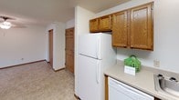 This single-floor residential unit offers a practical and comfortable layout designed for everyday living. The kitchen is well-appointed with warm oak cabinetry, light-toned countertops, and a full suite of white appliances including a coil-top range, refrigerator, dishwasher, and a double-basin stainless steel sink. Ample upper and lower cabinet storage makes the kitchen both functional and organized, with tile-style flooring adding easy-clean durability underfoot.

The open living and dining area is generously proportioned, with a seamless transition between tiled entryway flooring and plush wall-to-wall carpeting. Natural light filters in through a window fitted with vertical blinds, and a window-mounted air conditioning unit provides additional climate control. A central ceiling fixture illuminates the space, while a closet and entry door are conveniently accessible from this common area.

The bedrooms are comfortably sized, each featuring soft carpeting, textured ceilings, baseboard heating, and windows with vertical blinds that invite natural light while maintaining privacy. The full bathroom is clean and straightforward, featuring a wood-toned vanity with storage, a large mirror with overhead lighting, tile-style flooring, and a shower-tub combination with a curtain enclosure. Throughout the unit, consistent warm wood tones and neutral walls create a cohesive, understated aesthetic.