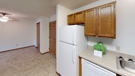 This single-floor residential unit offers a practical and comfortable layout designed for everyday living. The kitchen is well-appointed with warm oak cabinetry, light-toned countertops, and a full suite of white appliances including a coil-top range, refrigerator, dishwasher, and a double-basin stainless steel sink. Ample upper and lower cabinet storage makes the kitchen both functional and organized, with tile-style flooring adding easy-clean durability underfoot.

The open living and dining area is generously proportioned, with a seamless transition between tiled entryway flooring and plush wall-to-wall carpeting. Natural light filters in through a window fitted with vertical blinds, and a window-mounted air conditioning unit provides additional climate control. A central ceiling fixture illuminates the space, while a closet and entry door are conveniently accessible from this common area.

The bedrooms are comfortably sized, each featuring soft carpeting, textured ceilings, baseboard heating, and windows with vertical blinds that invite natural light while maintaining privacy. The full bathroom is clean and straightforward, featuring a wood-toned vanity with storage, a large mirror with overhead lighting, tile-style flooring, and a shower-tub combination with a curtain enclosure. Throughout the unit, consistent warm wood tones and neutral walls create a cohesive, understated aesthetic.