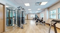 Located at 17783 Whitney Rd in Strongsville, Ohio, this fitness center offers a clean, modern environment designed to support a comprehensive workout experience. The single-floor space is organized to accommodate a diverse range of exercise equipment, including Matrix treadmills, elliptical machines, cable weight systems, adjustable benches, and multi-function strength training stations.

The interior features light hardwood-style flooring that adds warmth to the otherwise crisp, neutral aesthetic. A drop tile ceiling with recessed panel lighting ensures the space is consistently and evenly illuminated throughout. Multiple windows along the perimeter walls allow natural light to filter in, offering views of the surrounding outdoor landscape and contributing to an open, airy atmosphere.

Wall-mounted mirrors along one side of the room visually expand the space and provide users with real-time form feedback during exercise. A wall-mounted flat-screen television is also present, adding an entertainment option for cardio workouts. The layout allows for generous spacing between equipment, enabling comfortable movement and use of multiple stations simultaneously.

Glass-panel partitions and wooden doors provide defined entry and exit points, giving the space a polished, professional feel. Overall, this fitness center presents a bright, functional, and well-maintained environment suitable for a wide range of fitness activities.