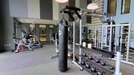 This contemporary fitness center is designed to accommodate a wide variety of workout styles and fitness goals. The space features a clean, modern aesthetic with a combination of neutral tones, textured wall panels, and large mirrors that enhance the sense of openness and allow users to monitor their form during exercise. The flooring consists of durable rubber gym matting, providing safety and comfort for high-impact activities. A standout feature of the facility is the heavy-duty TKO punching bag, suspended from a ceiling-mounted bracket, offering dedicated boxing and combat training capabilities alongside a pull-up bar station. The free weight section is well-stocked with a full rack of kettlebells in graduated sizes, catering to both beginners and advanced athletes. The cardio zone, visible in the background, is lined with a selection of machines including treadmills and upright exercise bikes, all neatly arranged for efficient use of the space. Additional functional training equipment, including weight plates and resistance accessories, is organized on nearby storage racks. The facility is well-lit with overhead pendant lighting, creating a bright and energizing workout environment. Overall, this fitness center is a comprehensive and thoughtfully appointed space suited for a full range of exercise routines.