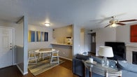 This single-story space in Arlington offers an open, functional layout that seamlessly connects the main living areas. The living room is anchored by a comfortable seating arrangement and features dark hardwood flooring that extends throughout the space, adding warmth and visual continuity. A side table with a lamp provides ambient lighting, complementing the overhead flush-mount ceiling light.

The dining area sits adjacent to the living room and accommodates a round white table with matching chairs, positioned atop a layered area rug. Colorful abstract wall art adds a vibrant accent against the neutral gray walls.

A pass-through counter connects the kitchen to the dining area, offering a convenient open feel while maintaining a defined cooking space. The kitchen features light upper cabinetry and a mirrored backsplash that reflects natural light into the interior.

A hallway visible from the main living area leads to additional rooms, with a white door and a secondary door visible at the end, suggesting a private bedroom and bathroom beyond. The textured ceiling and consistent gray wall tones create a cohesive, understated ambiance throughout the space.
