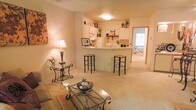 This single-floor residence offers an open-concept layout that seamlessly connects the living area to the kitchen, creating a cohesive and functional living space. The living room is carpeted and comfortably sized, providing ample room for seating arrangements centered around a glass-top coffee table with ornate metalwork detailing. A coordinating side table with a table lamp adds a warm, ambient glow that complements the overhead flush-mount ceiling light.

The kitchen features white cabinetry and a curved breakfast bar countertop, accompanied by two black metal bar stools with woven seats — an ideal spot for casual dining or entertaining. Open shelving above the counter provides additional display and storage space, while white appliances, including a refrigerator, complete the kitchen setup. The cabinetry and clean lines give the kitchen a tidy, organized appearance.

A hallway visible beyond the kitchen leads to additional rooms, with a glimpse of a carpeted bedroom bathed in natural light from a window. The overall ambiance of the space is warm and welcoming, with neutral tones throughout that create a calm, livable environment.