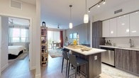 This thoughtfully designed single-floor residence combines modern aesthetics with functional living. The open-concept kitchen is a standout feature, boasting two-tone cabinetry — dark wood-grain lower cabinets paired with crisp white upper cabinets — complemented by quartz countertops, a full suite of stainless steel appliances, and a central island with bar seating. Pendant lighting and overhead track lighting illuminate the space effectively. Light wood-style flooring flows throughout, connecting the kitchen seamlessly to the living area. The living room is bright and welcoming, anchored by a ceiling fan, a large sliding glass door that opens to a private balcony, and views of the surrounding landscape. The bedroom features a distinctive warm-toned accent wall, a tray ceiling detail, ceiling fan, and ample natural light from a large window. A walk-in closet and access to a private ensuite are also present. The bathroom is finished with tile flooring, a dark wood-grain vanity, and a large mirror. A clean, light-filled entry hallway with light-toned flooring completes the layout. Throughout the residence, a neutral and contemporary color palette creates a cohesive, modern ambiance.