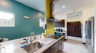 Located at 916 N College Ave in Bloomington, Indiana, this shared kitchen facility is thoughtfully designed to accommodate multiple users simultaneously across two distinct kitchen zones. The space makes a bold visual statement with its vivid teal and chartreuse accent walls, contrasted against rich dark-wood cabinetry and light-toned granite countertops that run throughout both areas.

The primary kitchen zone features a large central island with an integrated cooktop and a stainless steel range hood overhead, providing a functional and communal cooking hub. Ample storage is offered through floor-to-ceiling dark cabinetry, and the island's generous granite surface offers substantial workspace. A stainless steel refrigerator, double wall ovens, and a dishwasher round out the professional-grade appliance suite.

The secondary kitchen area mirrors the same design language, with open floating shelves mounted against the teal walls for easy access to cookware and supplies. Additional countertop space, a deep stainless steel sink, and a microwave make this zone fully self-sufficient. Natural light filters in through several windows, while recessed lighting ensures the space remains well-lit throughout. A wall-mounted television adds a casual, communal touch. The light tile flooring ties the two zones together, creating a cohesive and highly functional shared culinary environment.