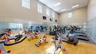 Located in Vista, CA, this fitness center offers a single, thoughtfully designed exercise room that provides a comprehensive workout environment. The space immediately impresses with its soaring ceiling height, which lends an open, airy feel, further enhanced by a row of clerestory windows positioned near the top of the walls that allow natural light to filter in throughout the day.

The flooring transitions between warm hardwood and durable rubber matting, delineating distinct workout zones within the room. A full wall of mirrors runs along one side, visually expanding the space and allowing users to monitor their form during exercise. Multiple flat-screen televisions are mounted at strategic heights around the room, providing entertainment during workouts.

The cardio zone features a lineup of treadmills, upright and recumbent stationary bikes, and spinning cycles, catering to a wide range of cardiovascular preferences. A dedicated strength training area includes a Vectra cable machine, a weight bench, and additional resistance equipment. The two-tone wall treatment — neutral beige above and cool slate blue below — gives the space a clean, modern aesthetic. Recessed ceiling lighting supplements the natural light, ensuring the room remains bright and welcoming at any hour.