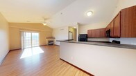 This single-floor residence offers an open-concept layout that seamlessly connects the kitchen and living areas, creating a bright and functional environment. The kitchen features rich, warm-toned cabinetry paired with dark countertops and a peninsula that defines the space while maintaining an open sightline into the living area. Black appliances, including a refrigerator, add a modern contrast to the cabinetry.

The living area is anchored by a fireplace, providing a natural focal point and adding warmth to the room. Vaulted ceilings enhance the sense of spaciousness, while a ceiling fan ensures year-round comfort. Light hardwood flooring flows throughout, tying the open-concept space together with a clean, cohesive look.

Natural light pours in through a large sliding glass door that opens to a balcony, offering outdoor access and views beyond. The neutral wall tones and simple trim work keep the overall aesthetic understated and versatile, making the space easy to personalize. The thoughtful layout and combination of functional kitchen design, comfortable living space, and outdoor access make this a well-rounded and inviting single-floor home.