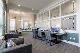 This single-floor community clubhouse offers a thoughtfully designed gathering space that blends comfort, functionality, and style. The interior is defined by a cohesive color palette of deep hunter green accent walls contrasted against crisp white trim and warm neutral tones, with crown molding adding a refined architectural touch throughout.

The main lounge area serves as the centerpiece of the space, featuring a dramatic stone fireplace flanked by dark built-in cabinetry and a mounted flat-screen television. Warm cognac leather sofas and accent chairs are arranged around a glass coffee table, creating an inviting setting for relaxation. A tray ceiling with recessed lighting enhances the sense of openness in this area. Adjacent to the lounge is a bar-height counter with leather barstools, ideal for casual gathering.

A separate recreation area accommodates a full-size billiards table beneath a modern pendant chandelier, with wall art and large windows that flood the room with natural light. Bold geometric metal wall décor adds a contemporary accent to the green feature wall.

The business center is equipped with multiple computer workstations along a long shared desk, offering a productive workspace within the clubhouse. Tile flooring transitions to plush carpet throughout the common areas, and a functional kitchenette with stainless steel appliances provides added convenience for residents and guests.
