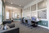 This single-floor community clubhouse offers a thoughtfully designed gathering space that blends comfort, functionality, and style. The interior is defined by a cohesive color palette of deep hunter green accent walls contrasted against crisp white trim and warm neutral tones, with crown molding adding a refined architectural touch throughout.

The main lounge area serves as the centerpiece of the space, featuring a dramatic stone fireplace flanked by dark built-in cabinetry and a mounted flat-screen television. Warm cognac leather sofas and accent chairs are arranged around a glass coffee table, creating an inviting setting for relaxation. A tray ceiling with recessed lighting enhances the sense of openness in this area. Adjacent to the lounge is a bar-height counter with leather barstools, ideal for casual gathering.

A separate recreation area accommodates a full-size billiards table beneath a modern pendant chandelier, with wall art and large windows that flood the room with natural light. Bold geometric metal wall décor adds a contemporary accent to the green feature wall.

The business center is equipped with multiple computer workstations along a long shared desk, offering a productive workspace within the clubhouse. Tile flooring transitions to plush carpet throughout the common areas, and a functional kitchenette with stainless steel appliances provides added convenience for residents and guests.