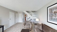 This two-storey space is designed with a functional and straightforward layout, centred around a bright, carpeted living area on the main floor. The living space features neutral grey walls and a textured popcorn ceiling, contributing to a clean and understated aesthetic. Plush, light-toned carpeting runs throughout, adding warmth underfoot and softening the overall atmosphere.

A prominent open staircase with white-painted wood railings connects the two floors, serving as a central architectural feature within the main living area. The staircase design allows sightlines to extend through the space, creating a sense of openness between levels. Adjacent to the staircase, a glimpse into what appears to be a kitchen area is visible, suggesting a practical and connected floor plan.

The main floor also includes multiple doorways, indicating additional rooms such as closets or utility spaces, contributing to the overall storage and functionality of the unit. Natural and artificial lighting work together to keep the space feeling bright and welcoming. The second floor, accessible via the central staircase, offers additional living or sleeping areas, completing the multi-level layout.