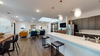 Located at 3845 Redan Rd in Decatur, Georgia, this single-floor communal space offers a welcoming and thoughtfully designed open-concept layout. Rich hardwood-style flooring flows throughout, tying together the various functional areas seamlessly.

The lounge area is anchored by bold, contemporary furnishings — a vibrant mustard yellow sofa and teal accent chairs with gold-toned throw pillows — set atop a graphic black-and-white striped area rug. A skylight overhead floods this central gathering zone with natural light, complemented by recessed lighting and decorative wall art that adds warmth and personality.

The kitchen is a standout feature, showcasing a generous white countertop island with bar seating for multiple guests, dark cabinetry with ample storage, and a full suite of stainless steel appliances including a refrigerator, built-in microwave, and double wall oven. Elegant pendant lights hang above the island, creating an inviting atmosphere for casual dining or socializing.

A dedicated dining area with a rustic wood high-top table and modern chairs sits near large windows, offering views of the lush greenery outside. Multiple exterior doors provide convenient access to the outdoor surroundings. Crown molding along the ceilings and crisp white walls throughout lend a clean, polished finish to the overall space.