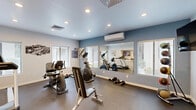 Located at 8429 N 27th Ave in Phoenix, Arizona, this dedicated fitness center offers a well-equipped exercise space designed to support a variety of workout routines. The room features a durable low-pile flooring surface suited for both cardio and strength training activities. Natural light filters in through multiple windows fitted with horizontal blinds, creating a bright and welcoming atmosphere, while recessed ceiling lights provide consistent illumination throughout. A wall-mounted mini-split air conditioning unit ensures a comfortable environment year-round — an essential feature in the Arizona climate. The exercise room is outfitted with a selection of cardio machines including a treadmill, elliptical trainer, and stationary bike, complemented by free weights, a weight rack, medicine balls, resistance bands, and a workout bench. A large wall-mounted mirror expands the sense of space and allows users to monitor their form during exercise. A wall-mounted television and posted workout charts add functional touches to the room. A pass-through window connects the fitness room to an adjacent space, adding openness to the layout. Motivational fitness-themed artwork adorns the walls, contributing to an energizing and purposeful ambiance throughout this thoughtfully arranged exercise facility.