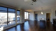 This single-floor space offers a thoughtfully designed open-concept layout that seamlessly connects the living, dining, and kitchen areas. Rich, dark hardwood flooring runs throughout, adding warmth and sophistication to the interior. The living area is bathed in natural light, courtesy of large floor-to-ceiling windows that frame expansive outdoor views and open directly onto a private balcony with metal railing — perfect for enjoying the surrounding scenery. A ceiling fan overhead enhances comfort and airflow in the main living space.

The kitchen is a standout feature, showcasing a central island topped with granite countertops, a sleek modern sink with a contemporary faucet, and dark cabinetry that complements the overall aesthetic. Pendant lighting hangs above both the kitchen island and the dining area, adding a refined, layered lighting scheme throughout the space.

Neutral-toned walls provide a clean, versatile backdrop that enhances the sense of openness. An elegant chandelier anchors the dining zone, while additional doorways suggest further private rooms beyond the open living area. The overall ambiance is modern, airy, and well-appointed, making this space both functional and visually striking.