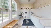 Located at 20 Graf Dairy Dr in Mobile, Alabama, the Pet Parlor at The Portier is a thoughtfully designed, single-room pet grooming facility that combines functionality with charming aesthetic details. The space features classic white subway tile wainscoting along the lower walls, transitioning to playful dog-print wallpaper above, creating a welcoming and thematic atmosphere for both pets and their owners.

The flooring consists of a black-and-white hexagonal mosaic tile pattern, adding a timeless, clean look that is both practical and visually appealing. A built-in tiled bathing tub occupies one wall, accompanied by a wall-mounted professional pet dryer with a flexible hose, making grooming tasks efficient and convenient. A dedicated grooming table with a grooming arm is positioned nearby, providing a proper workspace for styling and finishing.

Natural light fills the room through multiple windows, including a large double-hung window that offers views of the surrounding greenery, enhancing the bright and airy feel of the space. A glass-panel entry door provides additional light and easy access. A slatted wooden bench offers seating, while a dark utility table holds grooming supplies and accessories. The bold "PET PARLOR" signage mounted on the wall and the branded floor mat reinforce the space's identity as a premier pet care destination.