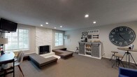 This single-floor space is thoughtfully arranged to serve multiple functions within an open, cohesive layout. The main gathering area is anchored by a floor-to-ceiling white stone fireplace, which serves as a striking focal point and lends a warm, rustic character to the room. Surrounding the fireplace, tiered upholstered platforms provide casual, flexible seating oriented toward a wall-mounted television, making the area well-suited for group gatherings or relaxed lounging.

Adjacent to the fireplace, a farmhouse-style coffee bar station features a gray sliding barn door cabinet stocked with shelving and storage, complemented by decorative signage and wall-mounted accents. A large oversized clock adorns the nearby wall, reinforcing the space's rustic-modern aesthetic. High-top stools and a bistro-style table nearby offer additional informal seating.

The reception or lounge zone near the entrance includes a pair of upholstered navy blue accent chairs, a small side table, and a dedicated workspace desk with white chairs, suitable for meetings or administrative tasks. A separate, partially visible office area with a desk and computer provides additional workspace.

Throughout, neutral gray carpet, recessed lighting, and light gray walls create a calm, unified atmosphere. Windows along the exterior walls allow natural light to filter in, enhancing the overall sense of openness and comfort.